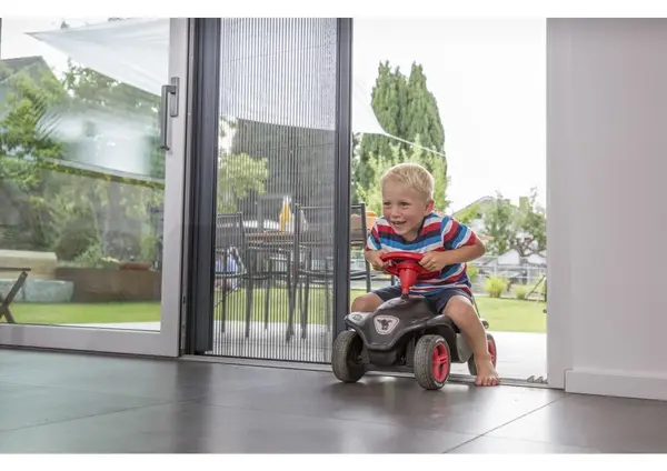 Ein fröhlicher Junge fährt auf einem Rutschauto durch eine offene Terrassentür mit Insektenschutz.