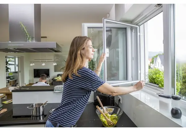 Frau lüftet Küche durch geöffnetes Fenster, frische Luft und Pflanzen am Fensterbrett.